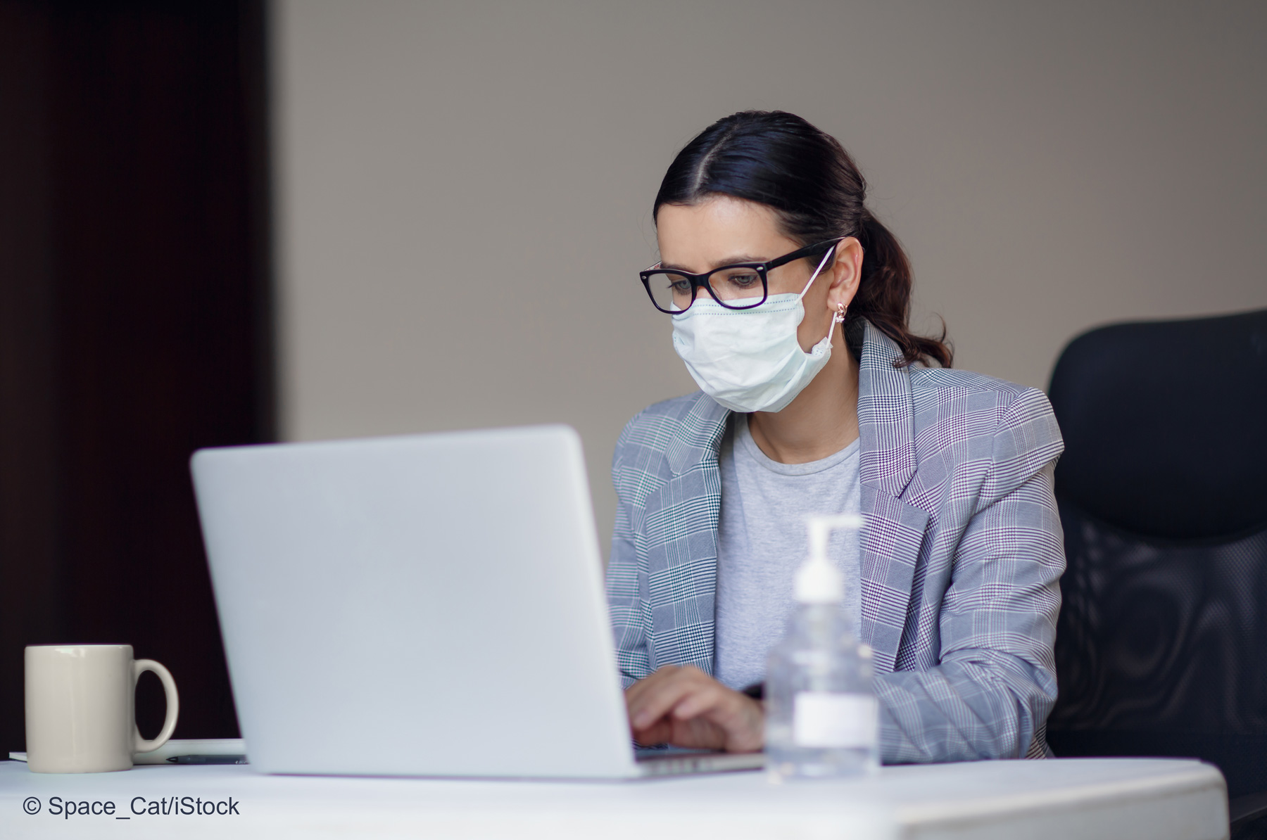 A woman wearing glasses and a face mask is focused on her laptop while working at a desk. A small mug and a bottle of hand sanitizer are on the table. The setting suggests a professional environment, emphasizing safety and health precautions.