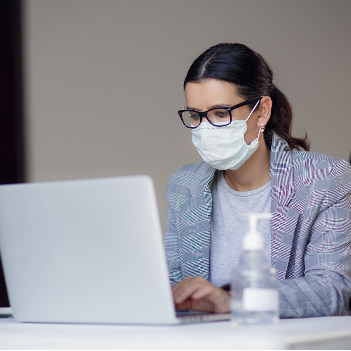 A woman in a mask and glasses is working on a laptop at a table. She is wearing a checkered blazer and appears focused on her task. A bottle of hand sanitizer is visible on the table next to her.