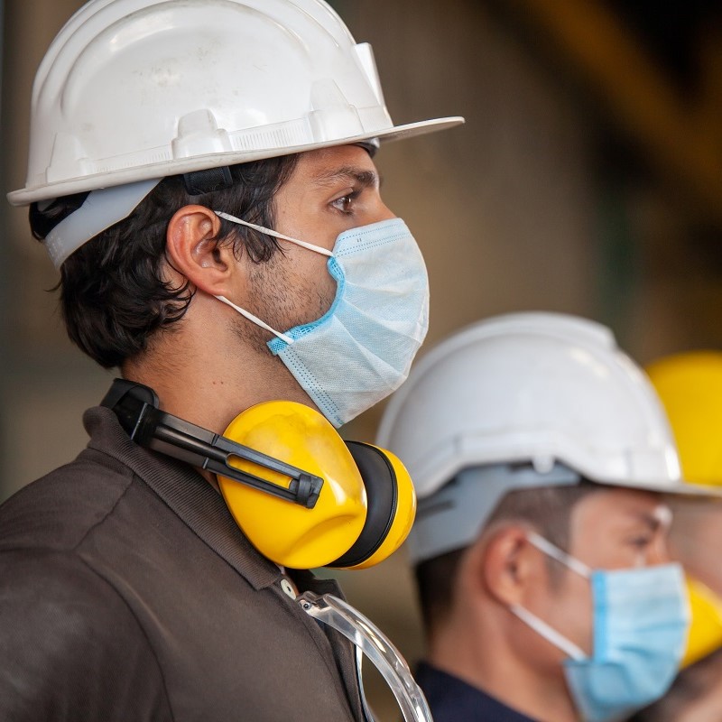 Two workers in safety gear are standing side by side. One has a white hard hat, a blue surgical mask, and yellow ear protection resting around his neck. The second worker also wears a hard hat and a mask. Their expressions suggest focus and readiness in a work environment.