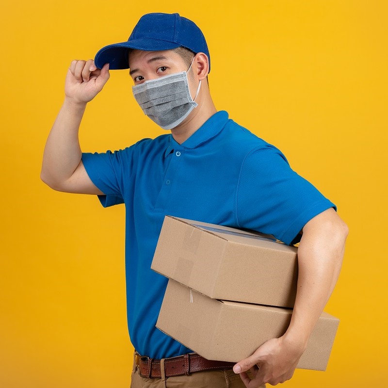 A delivery person wearing a blue polo shirt and a blue cap is standing against a yellow background. He is holding two cardboard boxes in one arm while tipping his cap with the other hand. He is also wearing a gray mask.