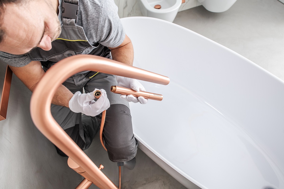 A plumber is working on a modern bathtub, carefully handling copper plumbing components and tools while wearing gloves. The bathtub is white and has a sleek design, set against a minimalist bathroom backdrop.