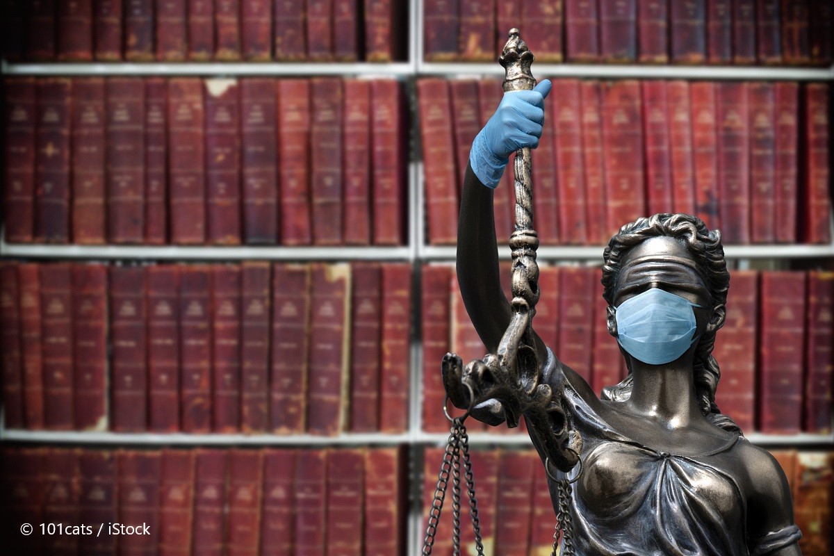 A statue of Lady Justice wearing a blue face mask and blue gloves holds a set of scales. She stands in front of a blurred background of stacked red books, symbolizing law and justice during the pandemic.