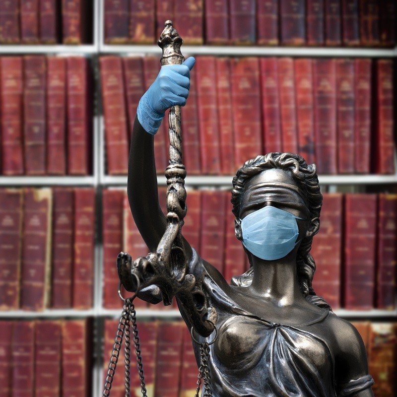 A statue of Lady Justice wearing a light blue face mask and blue gloves stands confidently, holding a scale in one hand. Behind her is a background of red-bound law books. The image symbolizes the ongoing themes of justice and public health.