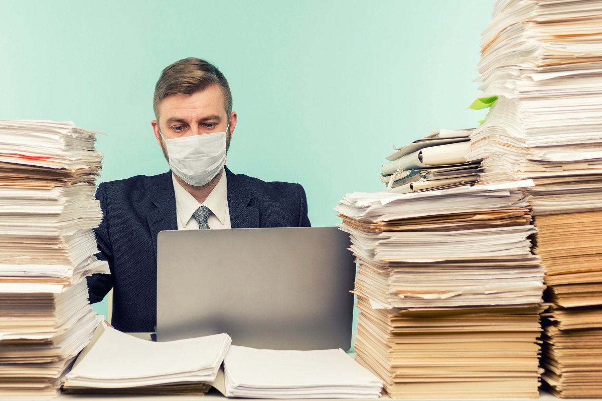 A man in a suit and face mask sits at a desk, focused on a laptop. He is surrounded by tall stacks of paper documents, indicating a heavy workload. The background is a light blue color.
