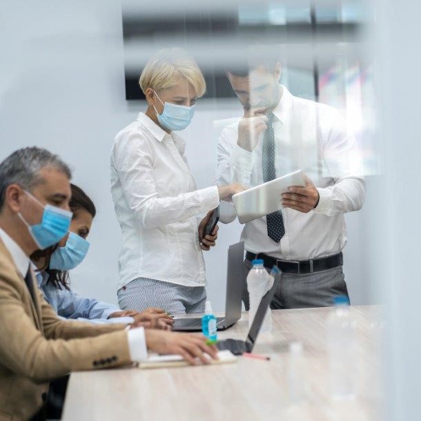A group of five people in a conference room are engaged in discussion while wearing face masks. Two individuals, a man and a woman, are collaborating over a tablet, while the others are focused on laptops at the table. A water bottle and hand sanitizer are also present.