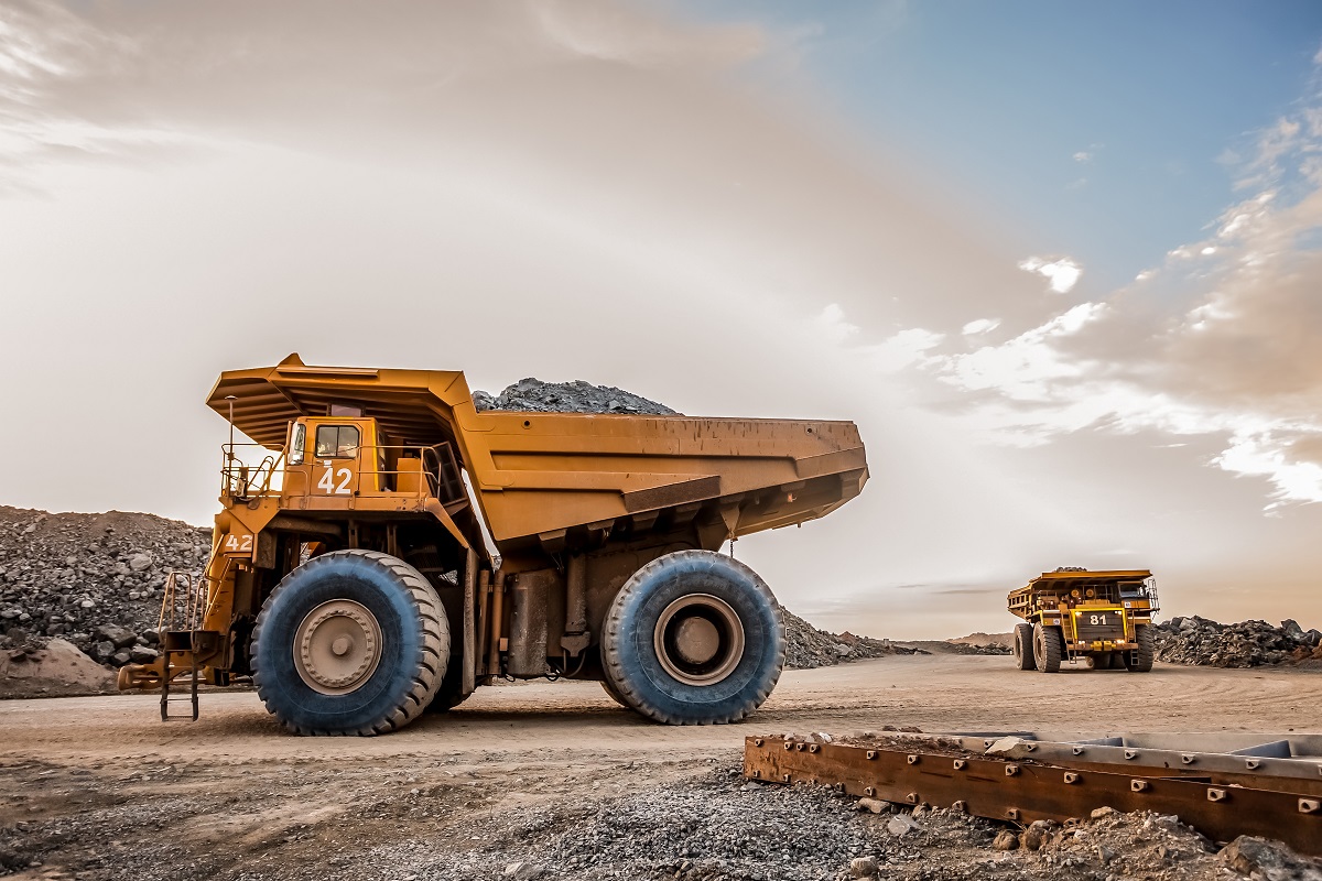 Two large yellow mining trucks are parked on a dirt road near a heap of materials. One truck is closer to the viewer, displaying the number 42, while a second truck, further back, has the number 81 visible. The sky is clear with subtle colors.