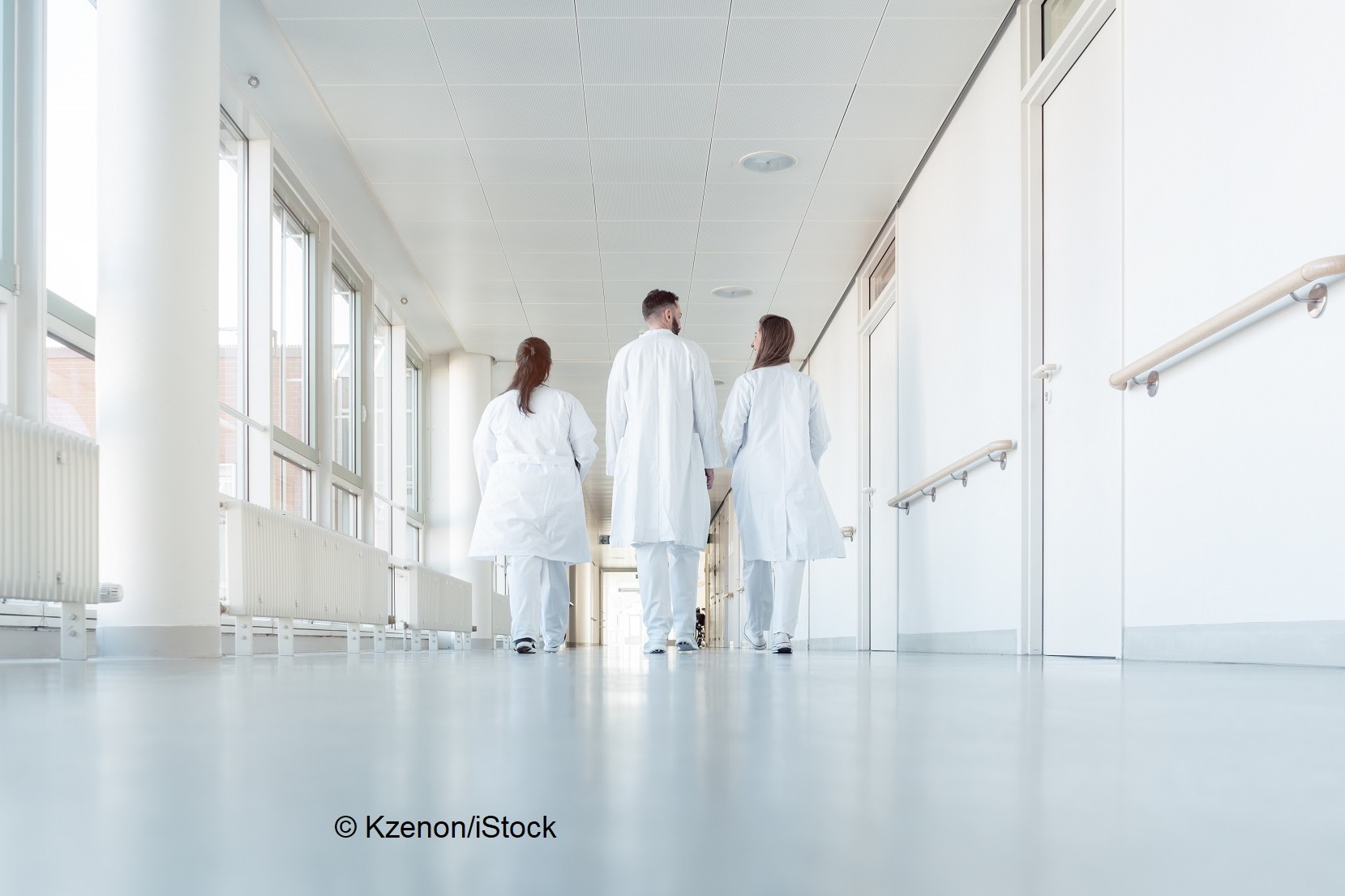 Three medical professionals in white lab coats walk together down a bright, modern hospital corridor. Large windows line the walls, allowing natural light to fill the space. The atmosphere is clean and clinical, emphasizing a focus on healthcare.
