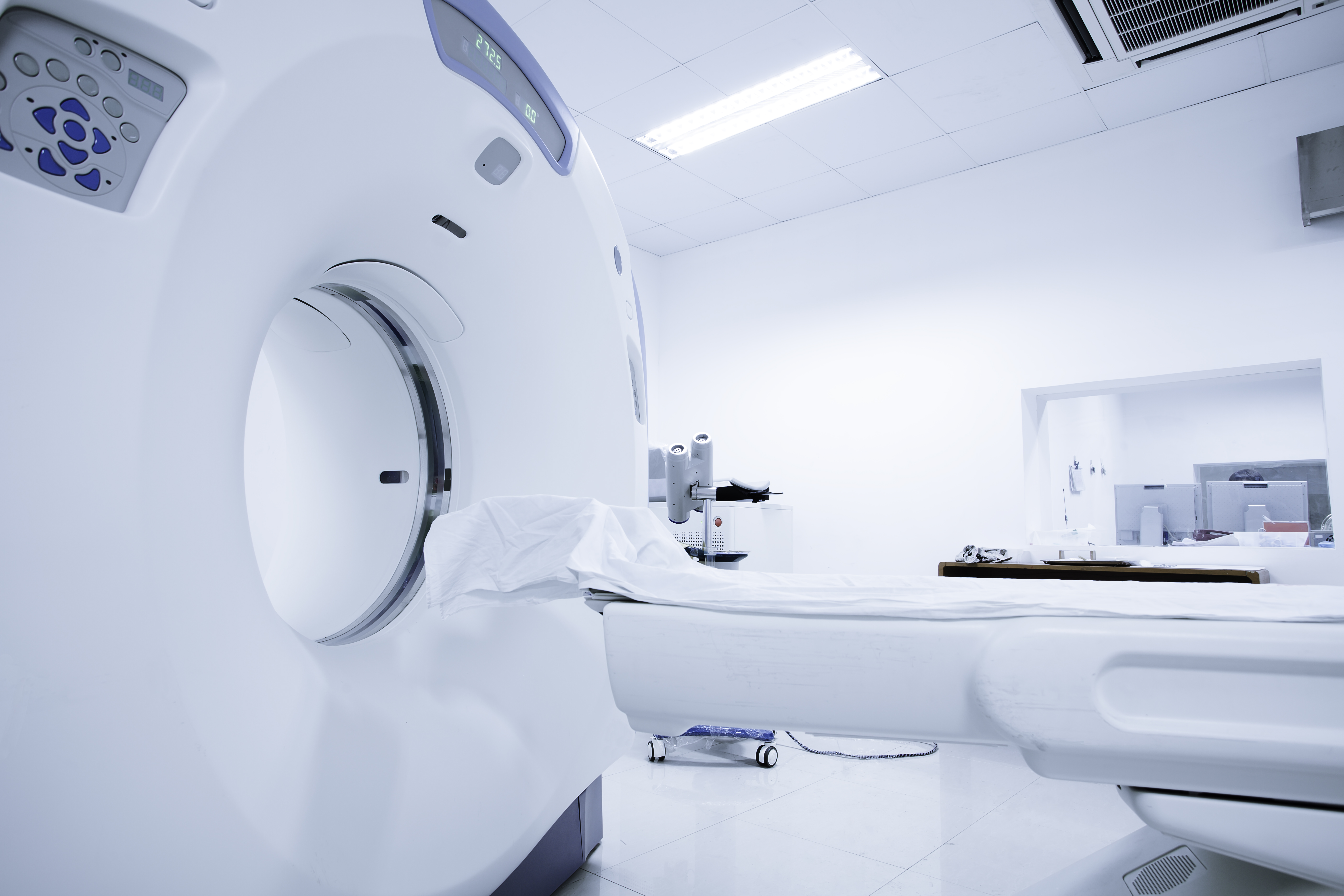 A CT scan machine is positioned in a bright, sterile room. A padded table, covered with a white sheet, extends towards the machine’s entrance. The environment appears clean and clinical, designed for medical imaging purposes.