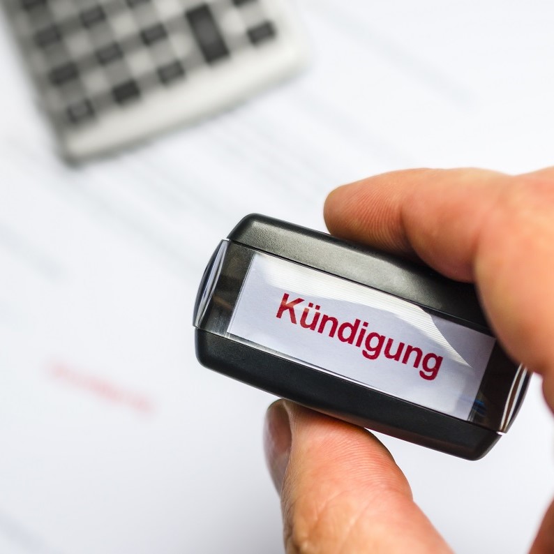 A hand holds a stamp labeled "Kündigung," which means "termination" in German. The background includes a blurred calculator and a sheet of paper, suggesting a context related to official documents or financial matters.