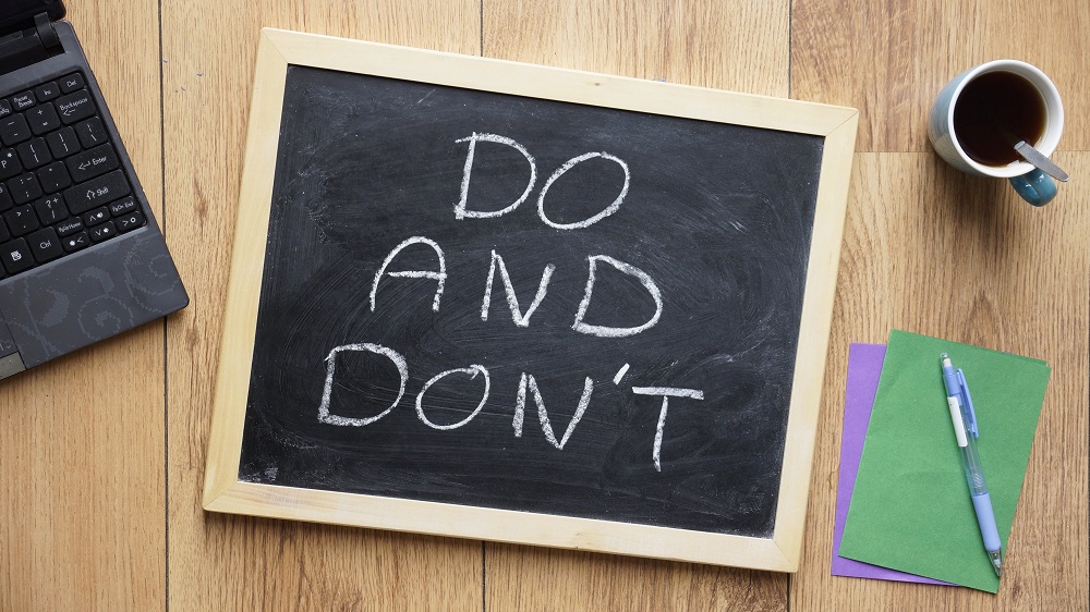A chalkboard with the words "DO AND DON'T" written in white chalk. The board is placed on a wooden surface, alongside a laptop keyboard, a cup of tea with a spoon, and some colorful folders.