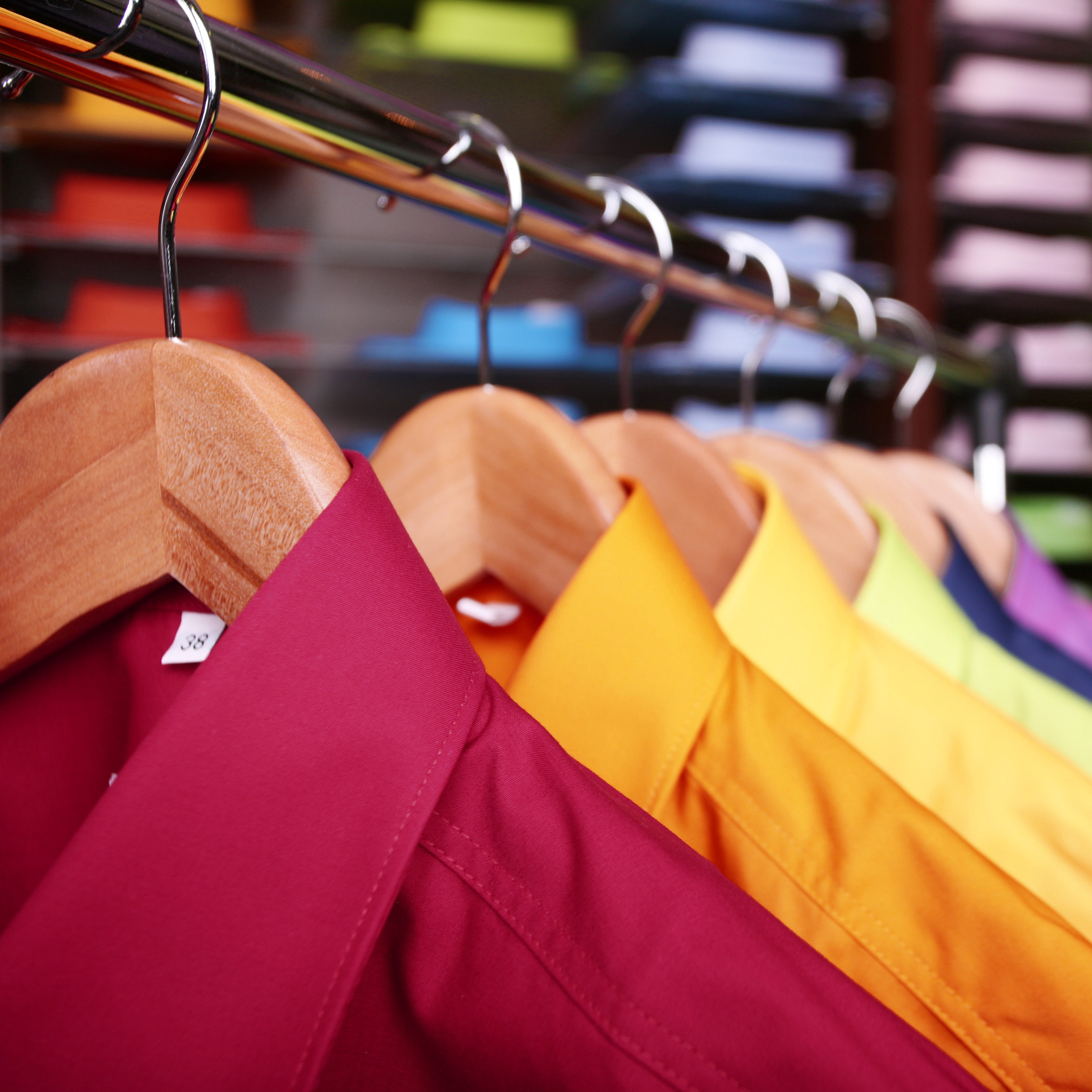 Auf einem Kleiderständer hängen mehrere Hemden in verschiedenen Farben. Die ersten beiden sind in einem dunklen Rot und leuchtenden Gelb, gefolgt von weiteren Hemden in anderen bunten Farbtönen. Der Hintergrund zeigt verschwommen stapelbare, farbige Materialien.