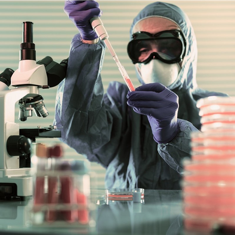 A person in protective gear, including gloves and a face mask, is holding a pipette filled with a red liquid. They are working at a laboratory table with a microscope and petri dishes in the background, focusing on a scientific experiment.