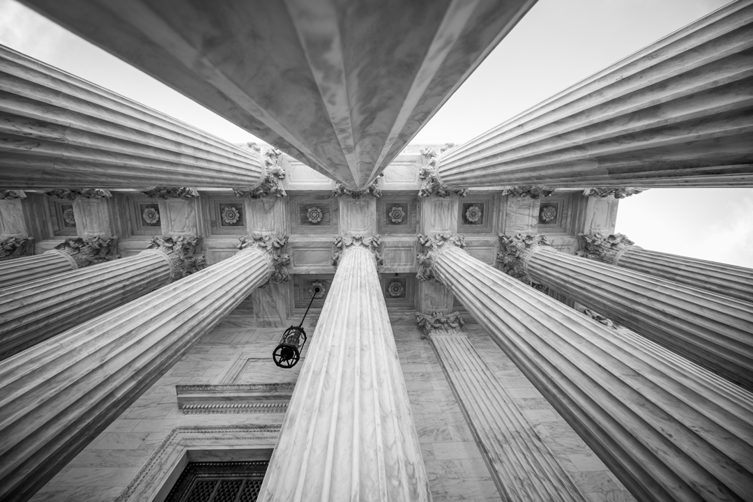 A low-angle view of grand, classical columns that support a decorative ceiling. The image is in black and white, highlighting the intricate details of the pillars and architectural elements around them. The atmosphere conveys a sense of historical significance and grandeur.