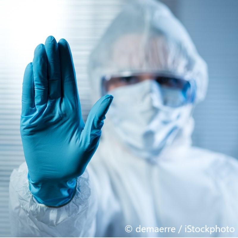 A person in protective clothing and gloves extends their hand forward in a "stop" gesture. The background is softly blurred, emphasizing the seriousness of the stance. The image conveys a message of caution and safety, likely in a medical or hazardous environment.