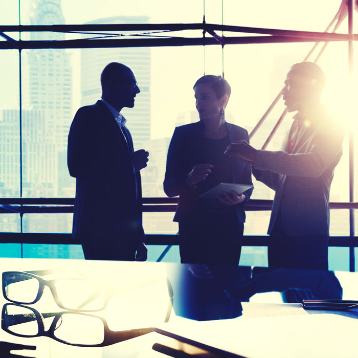 Drei Personen stehen in einem modernen Büro mit großen Fenstern. Sie diskutieren angeregt und halten dabei Notizen oder ein Tablet in der Hand. Im Vordergrund liegen eine Brille und einige Büromaterialien. Die Szene vermittelt eine geschäftliche Atmosphäre.