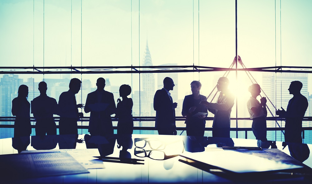 Silhouetten von sechs Personen stehen in einem modernen Büro mit einer großen Fensterfront. Sie scheinen in ein Gespräch vertieft zu sein, während die Sonne am Horizont aufgeht und die Skyline einer Stadt im Hintergrund sichtbar ist.