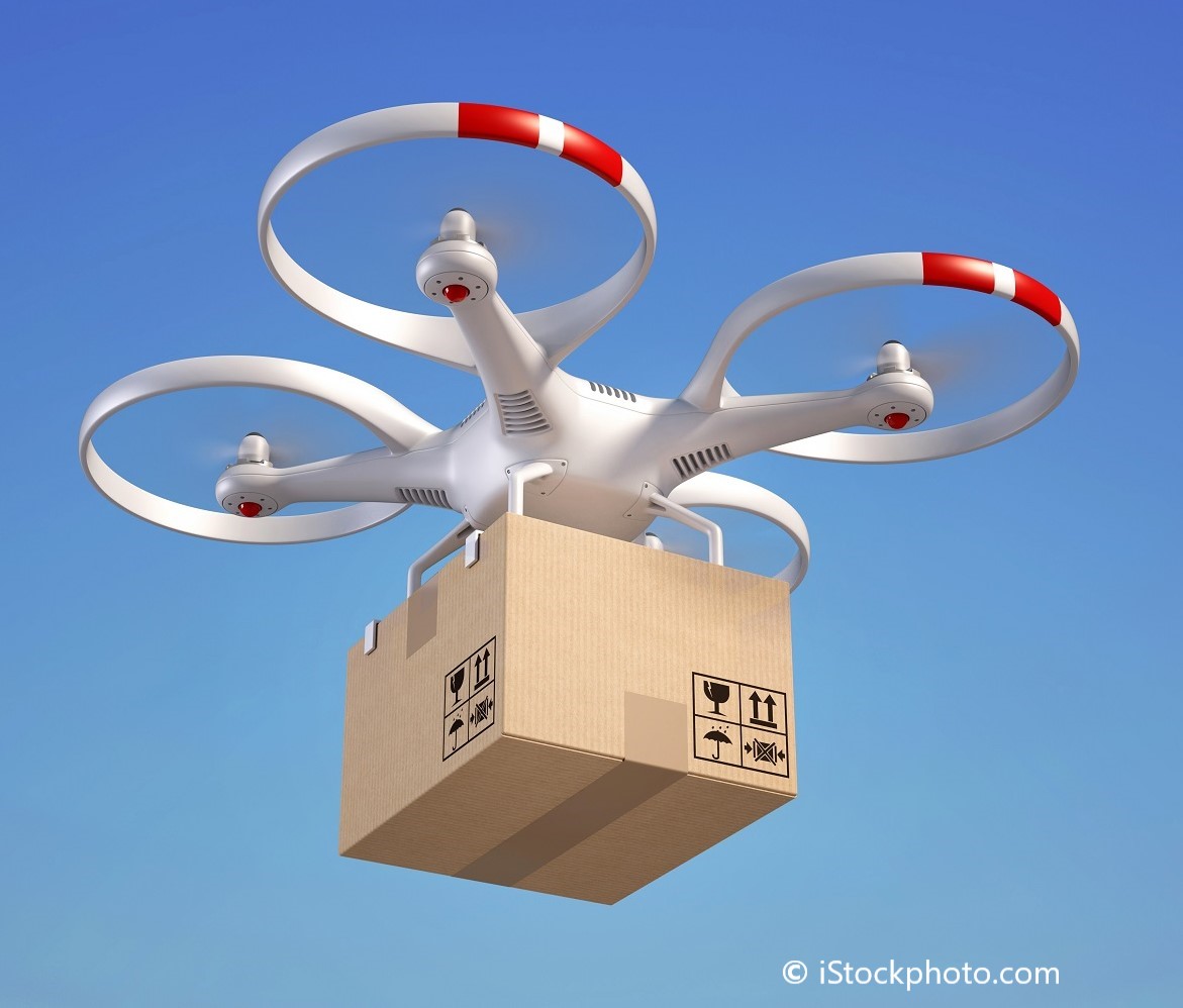 A white drone with red accents is flying against a blue sky, carrying a cardboard box. The box features safety icons indicating fragile contents. The image highlights the concept of drone delivery services.