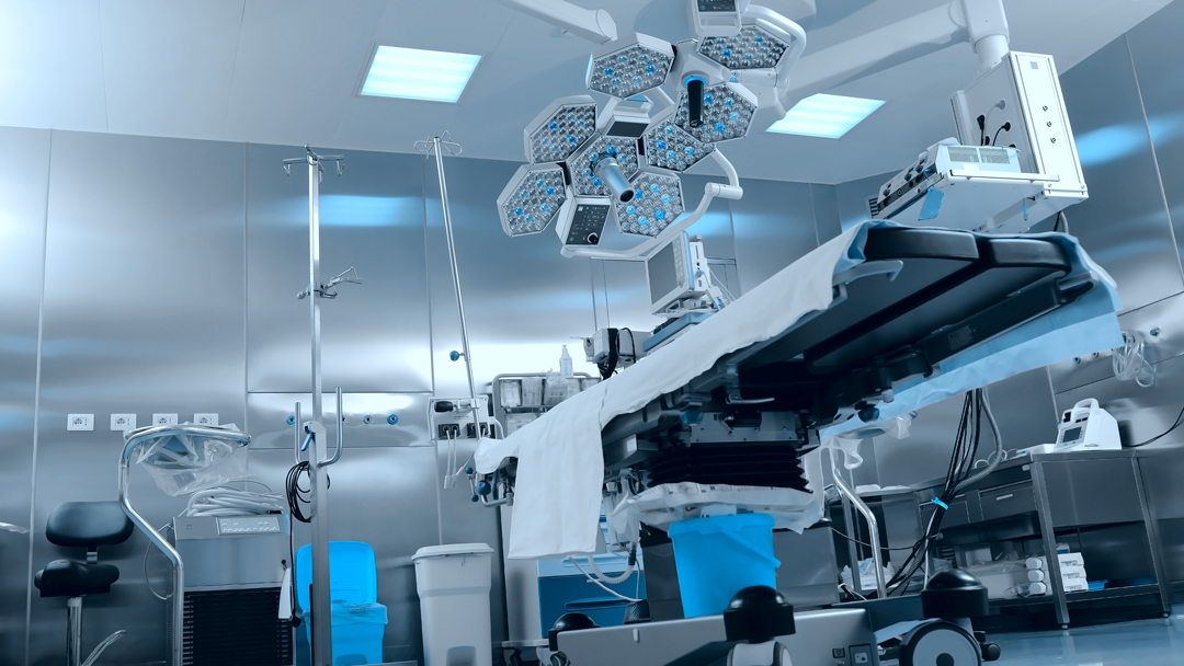 Ein modernes Operationszimmer mit einem chirurgischen Tisch in der Mitte, der mit einem weißen Laken bedeckt ist. Über dem Tisch hängen mehrere chirurgische Lampen. Im Hintergrund sind medizinische Geräte und eine Infusionsstation sichtbar. Die Wände sind metallisch und reflektierend.