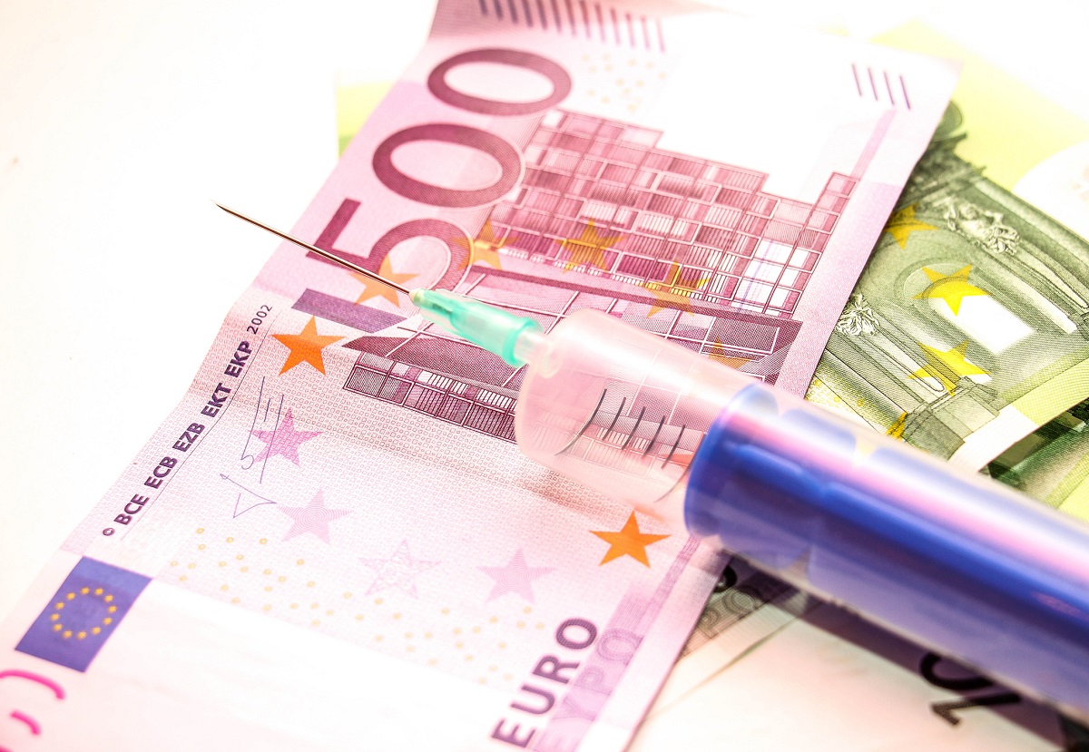 Eine Spritze liegt auf einem Stapel von Euro-Banknoten, darunter ein 500-Euro-Schein. Die Spritze hebt sich durch ihre transparente Kappe und die grüne Nadel ab. Die Banknoten sind teilweise sichtbar und deuten auf finanzielle Themen hin.
