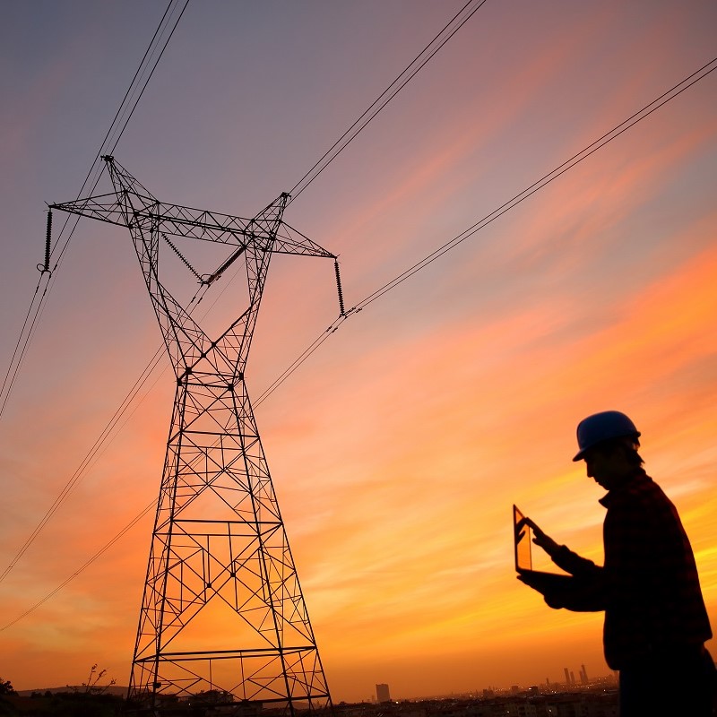 A silhouetted worker wearing a hard hat stands with a tablet in front of a tall power transmission tower against a vibrant sunset sky, showcasing the intersection of technology and energy infrastructure.