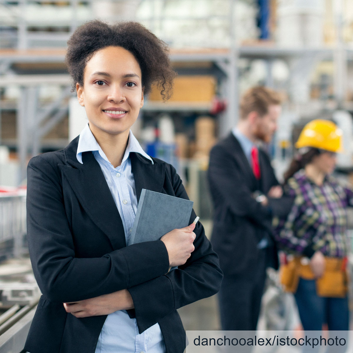 A confident woman in a black blazer stands in a warehouse, holding a notebook. In the background, two colleagues are engaged in conversation, with one wearing a hard hat. The setting suggests a professional industrial environment.