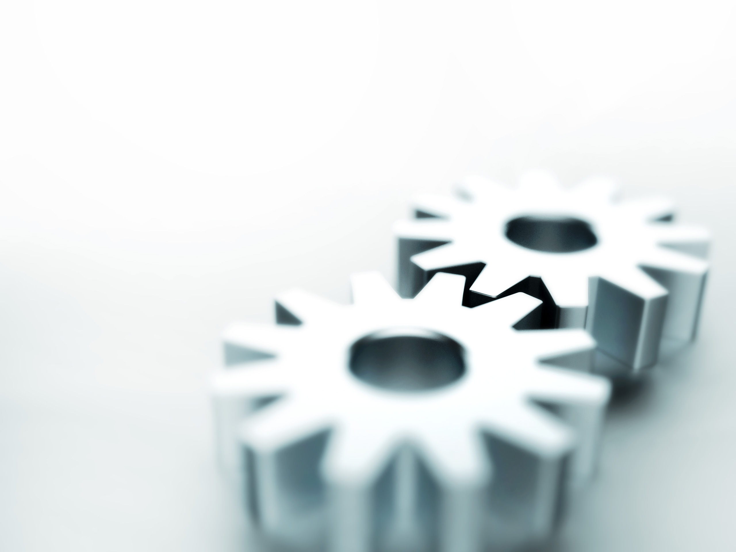 Two metallic gears are positioned close together on a light background. The gears have a star shape with central holes, suggesting a mechanical theme. The image is slightly blurred, creating a soft focus effect.