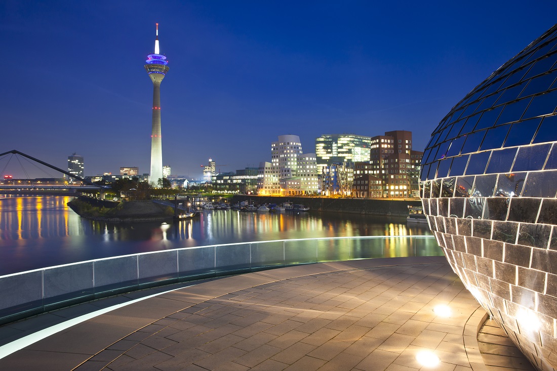 Eine Nachtaufnahme von Düsseldorf zeigt den Fernsehturm mit bunten Lichtern, umgeben von modernen Gebäuden und einem Fluss. Im Vordergrund ist ein runder, gläserner Bau zu sehen. Die Stadt ist beleuchtet und reflektiert auf der Wasseroberfläche.