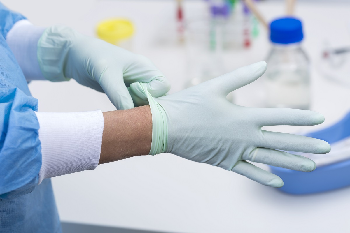 A person is putting on a light green latex glove, preparing for a medical procedure. The scene suggests a sterile environment, with blurred laboratory equipment in the background.