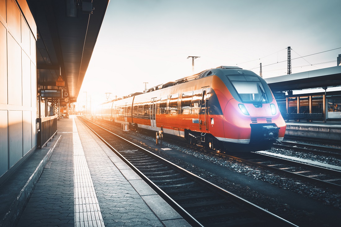A modern train with a sleek design is stationary on railway tracks at a station during sunset. The scene is illuminated by warm light, highlighting the train’s bright colors. The platform is partly visible, along with the tracks extending into the distance.