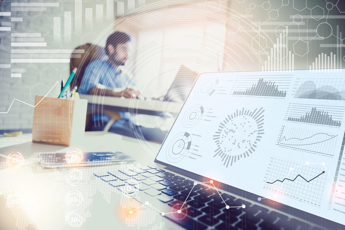 A laptop displays various data visualizations and charts, including bar graphs and pie charts, indicating analysis and metrics. A smartphone rests nearby, while a person is seen working in the background, suggesting a busy office environment focused on data-driven tasks.