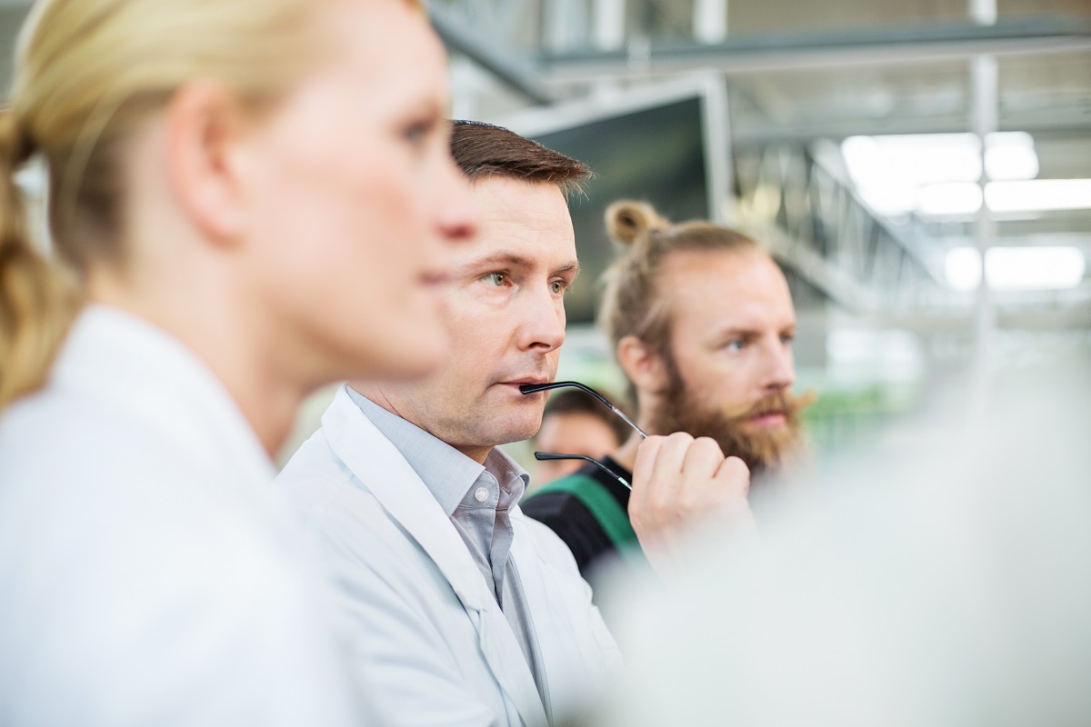 Eine Gruppe von drei Personen in weißen Laborkitteln schaut aufmerksam in eine Richtung. Einer der Männer hält eine Brille in der Hand und wirkt nachdenklich. Der Hintergrund ist unscharf, was die Konzentration auf die diskutierende Gruppe lenkt.