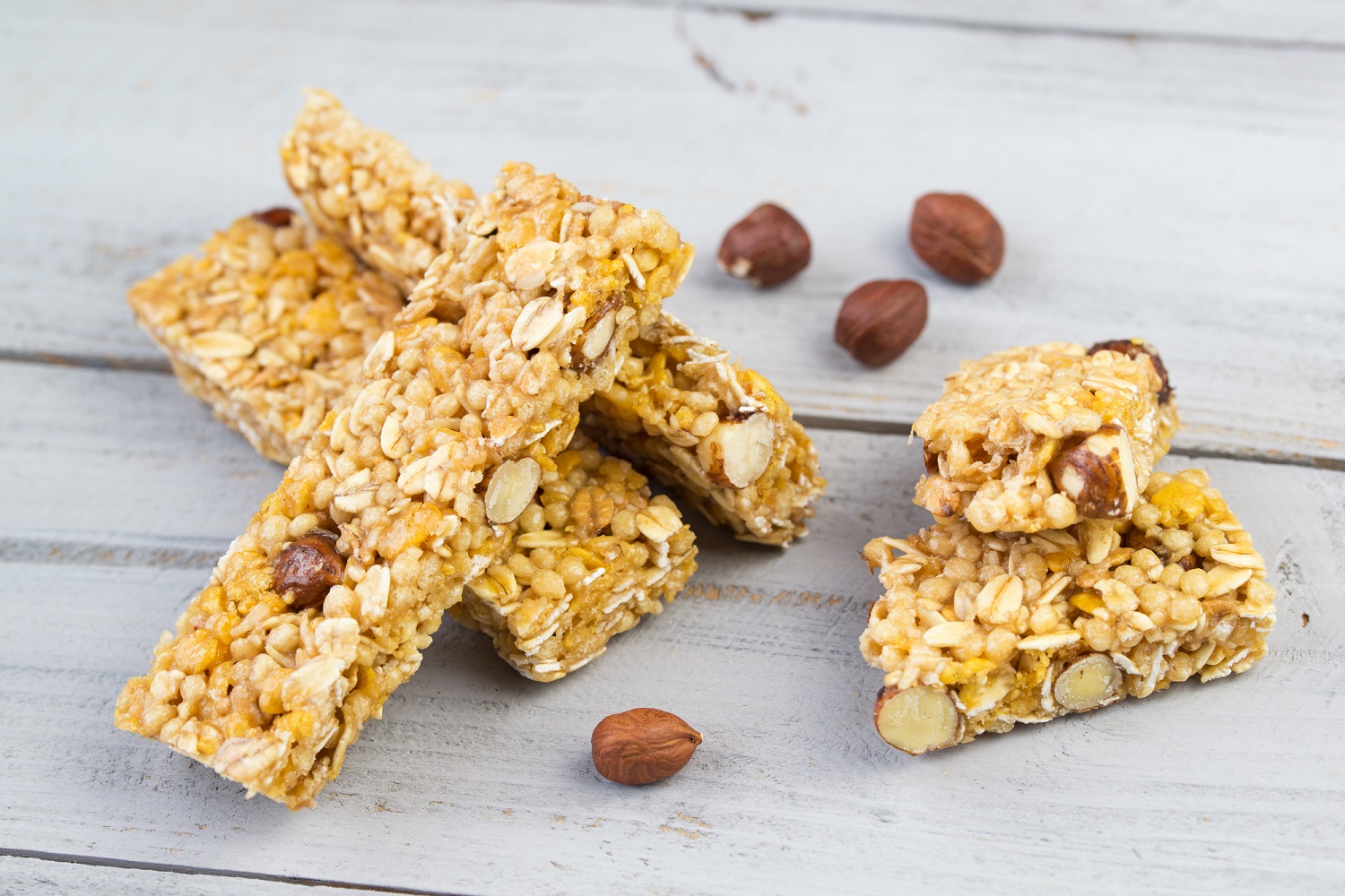 Three granola bars made with oats and nuts are arranged on a light wooden surface. A few whole hazelnuts are scattered nearby. The bars appear crunchy and are held together by a golden syrup, suggesting a healthy snack option.