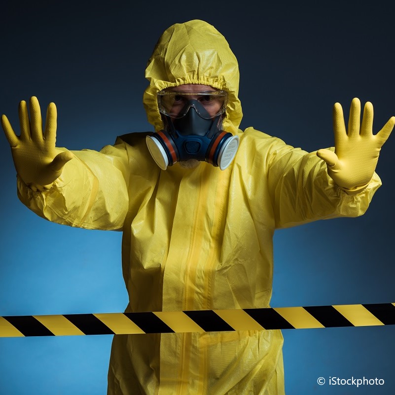 A person wearing a yellow protective suit, gloves, and a gas mask stands with their hands raised in a stopping gesture. A caution tape is visible in front, indicating a hazardous area. The background is softly lit in shades of blue.