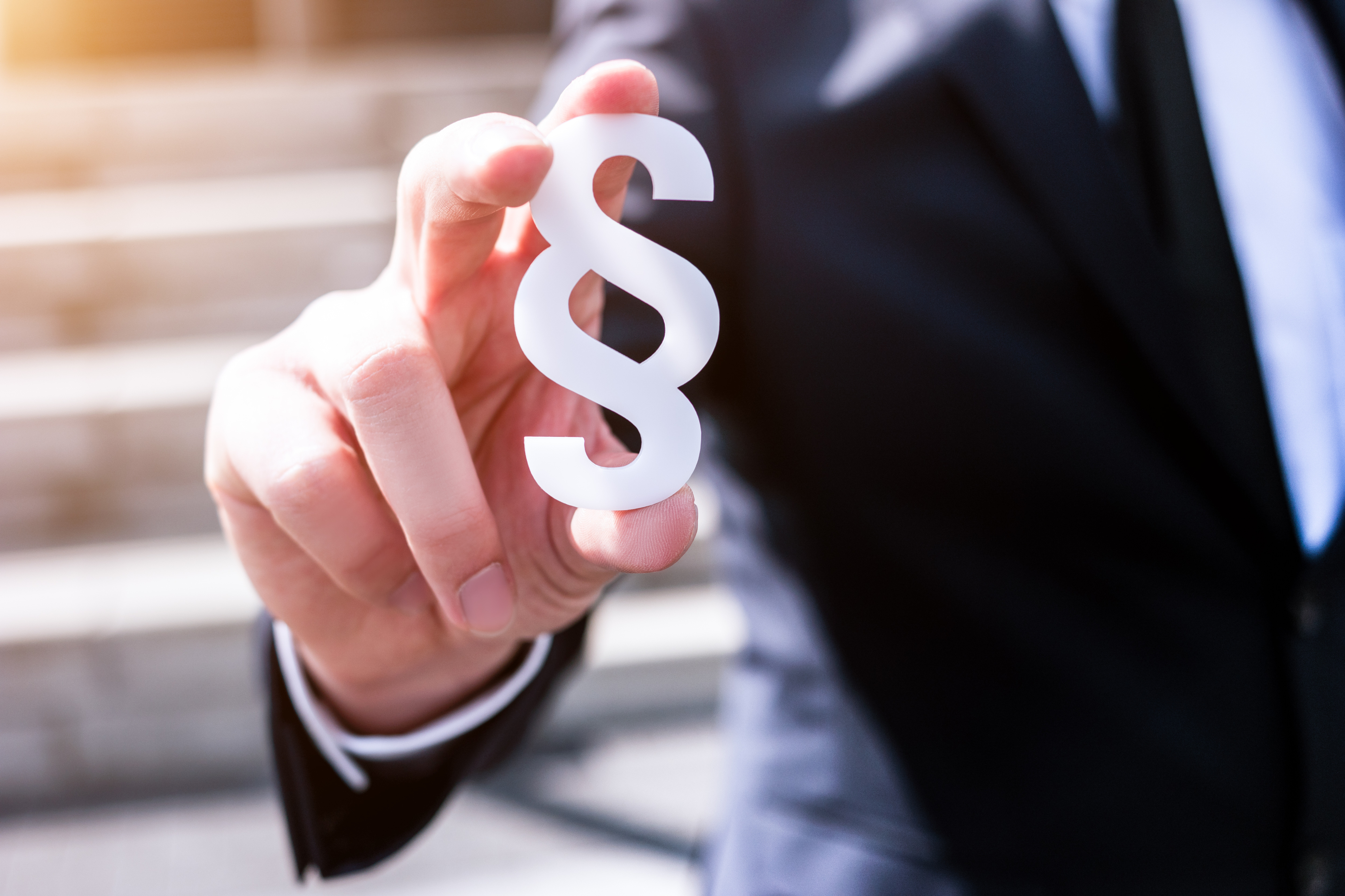 A person in a dark suit is holding a stylized paragraph symbol with their right hand. The background is blurred, featuring steps and a soft light, emphasizing the importance of legal representation or regulations.