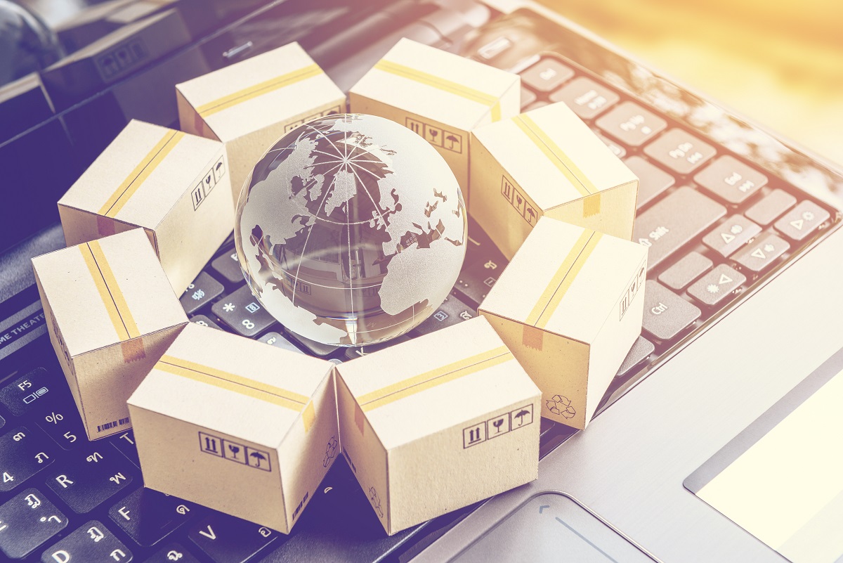 A transparent globe sits at the center of a circle formed by small cardboard boxes. The background features a laptop keyboard, symbolizing e-commerce and global shipping. The boxes, marked with tracking symbols, represent packaged goods ready for delivery.