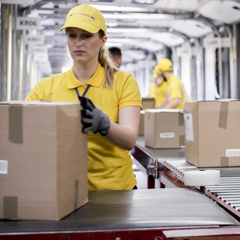 A woman in a yellow shirt and cap is working in a packaging area, handling a cardboard box. She is focused on her task, with other workers visible in the background, also involved in processing packages.
