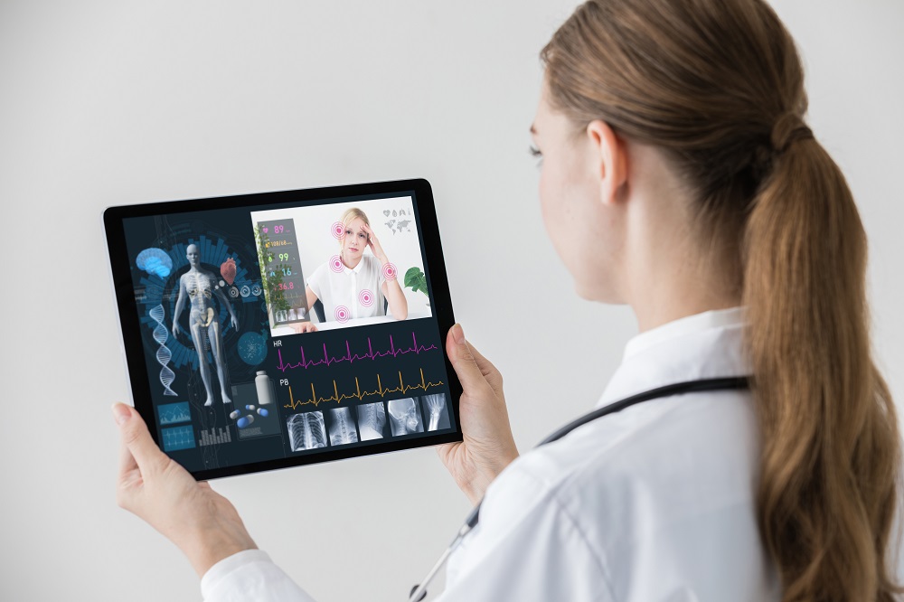 A healthcare professional holds a tablet displaying a virtual consultation. The screen shows a remote patient expressing discomfort, with visual data representing vital signs and a human anatomy graphic. The setting suggests a focus on telemedicine and patient assessment.