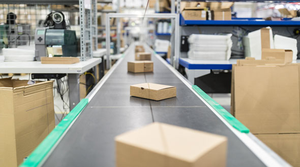 A conveyor belt in a warehouse carries multiple cardboard boxes. The background shows shelves stocked with more packaging materials and supplies, creating a bustling environment focused on order fulfillment and logistics.