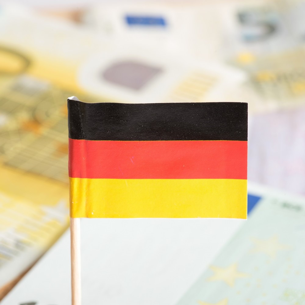 A small flag of Germany with three horizontal stripes: black on the top, red in the middle, and yellow at the bottom. The flag is positioned against a blurred background of Euro banknotes.