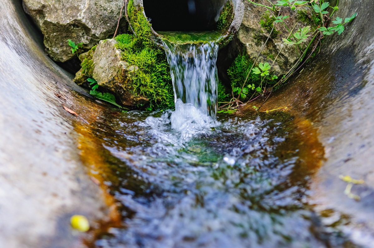 A small stream of clear water flows from a concrete pipe surrounded by mossy rocks. The water ripples as it glides over a surface, reflecting natural greenery nearby.
