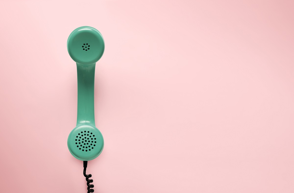 A vintage green telephone receiver hangs against a soft pink background. The receiver has a spiral cord attached, emphasizing its retro style. The image conveys a sense of nostalgia and communication.