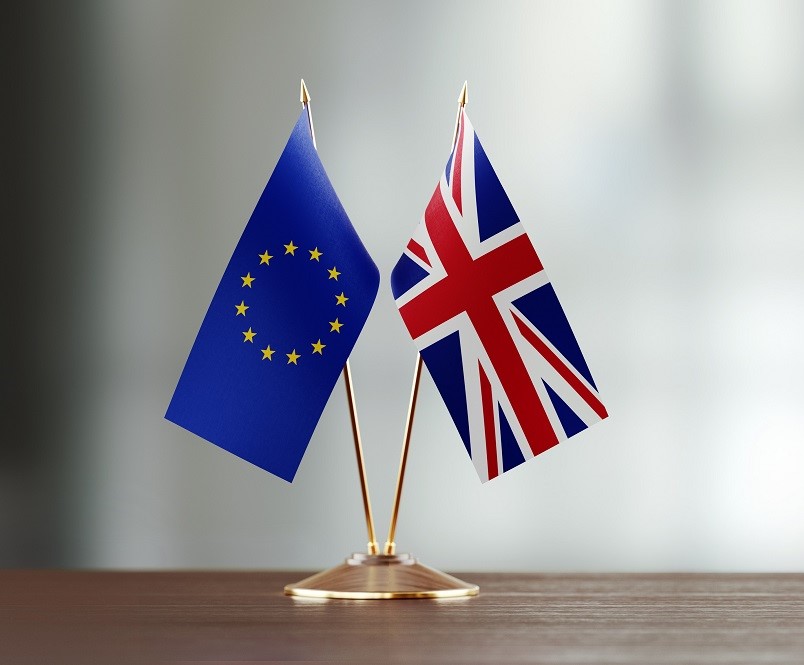 Two small flags are displayed on a wooden surface. One flag represents the European Union with a blue background and yellow stars in a circle. The other flag represents the United Kingdom with a red, white, and blue design featuring a Union Jack pattern.