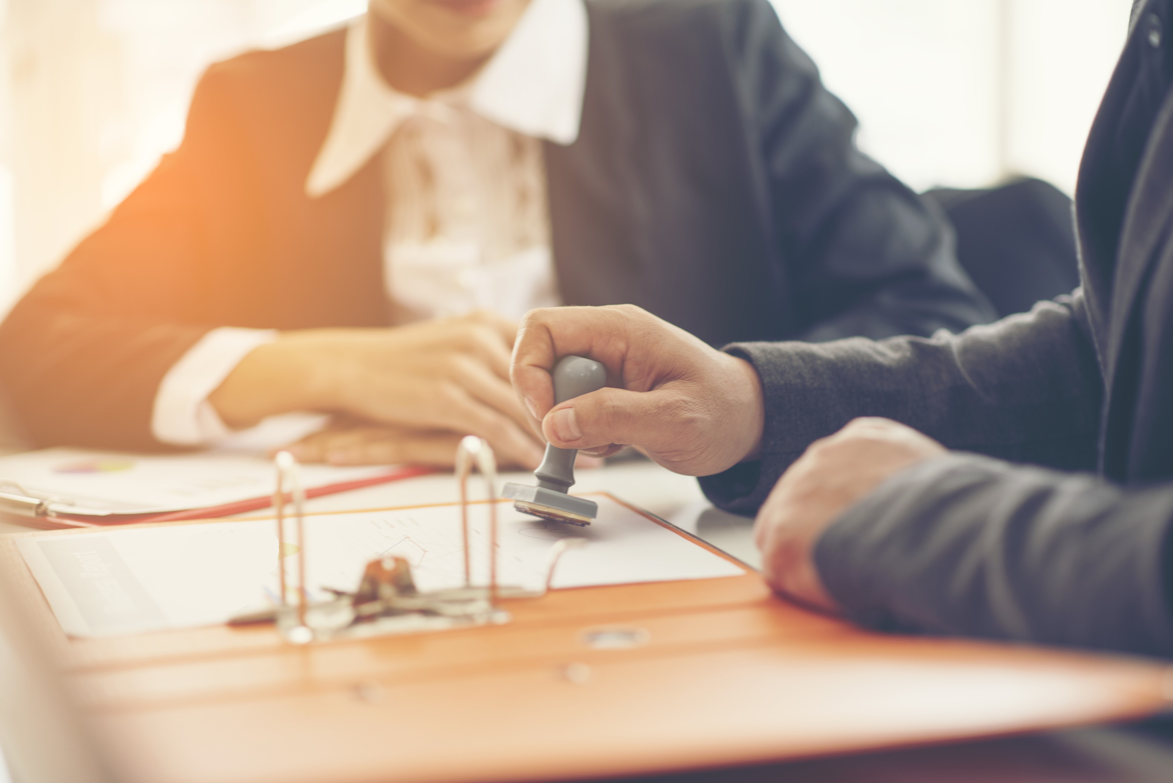 A hand is applying a stamp to a document on an orange binder, while another person is seated nearby, partially visible. The setting suggests a business meeting or official signing process.