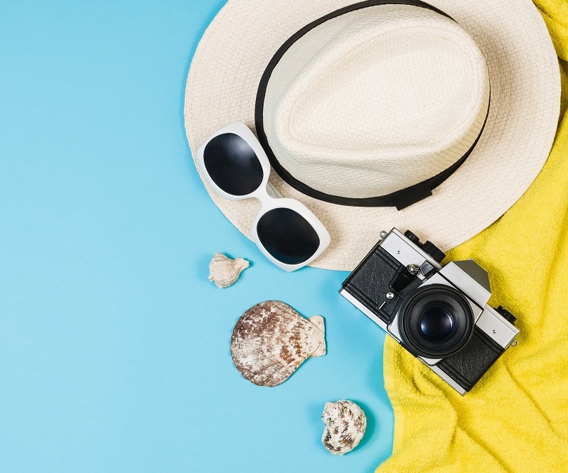 The image features a straw hat, black sunglasses, and a vintage camera arranged on a bright blue background. Shells are scattered nearby, with a folded yellow towel partially visible. The overall theme suggests a relaxing beach or travel experience.