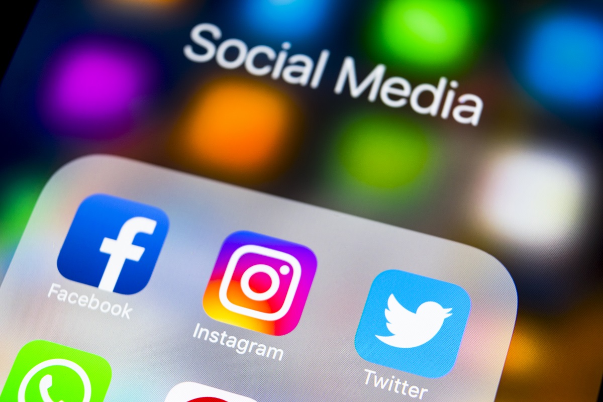A close-up image of a smartphone screen displaying various social media app icons, including Facebook, Instagram, and Twitter, along with the text "Social Media" at the top. The background features colorful blurs and light effects.