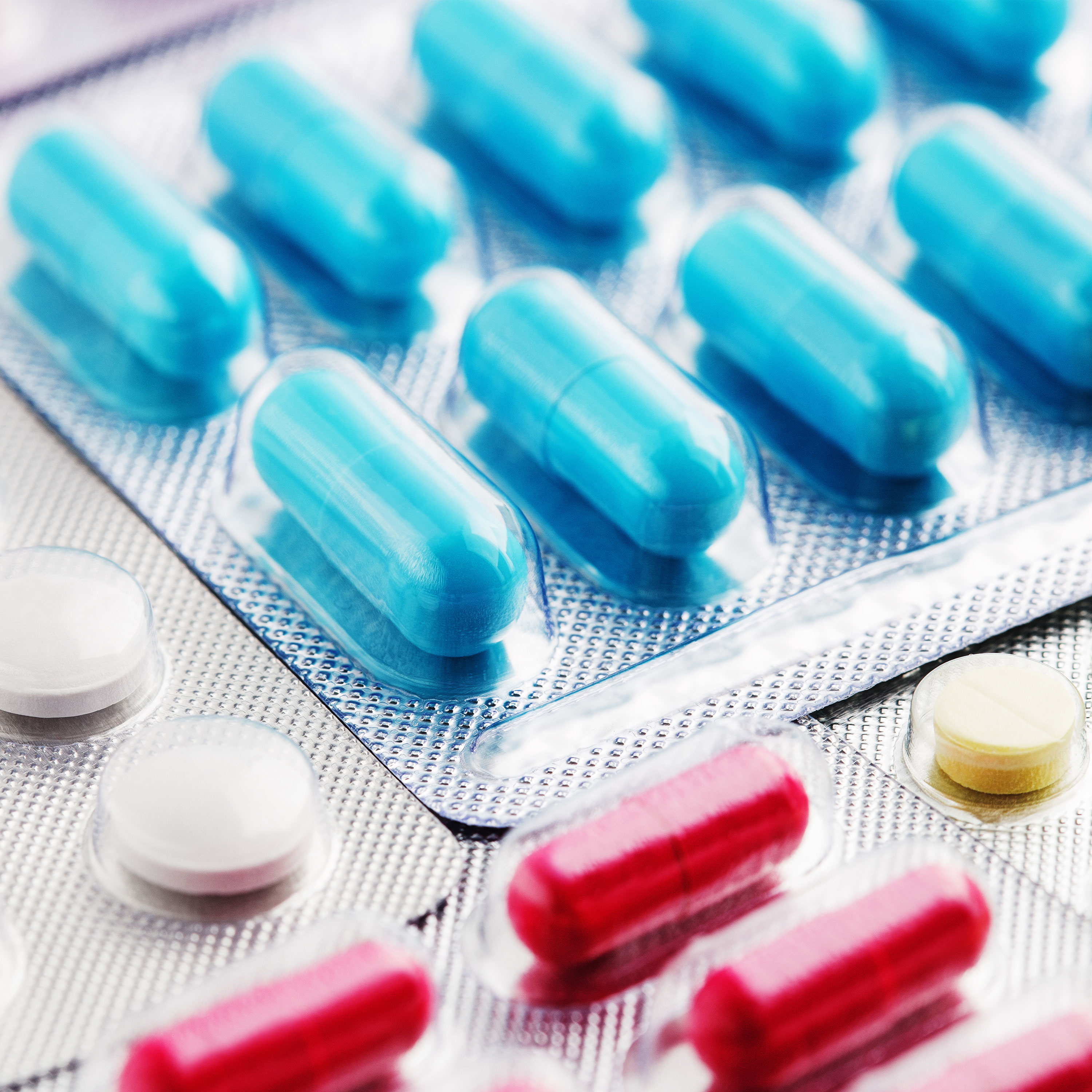 A close-up image of multiple blister packs containing various capsules and tablets. Prominent colors include blue capsules, red capsules, as well as white and yellow tablets, arranged neatly within their packaging. The focus is on the medication, highlighting diversity in shapes and colors.