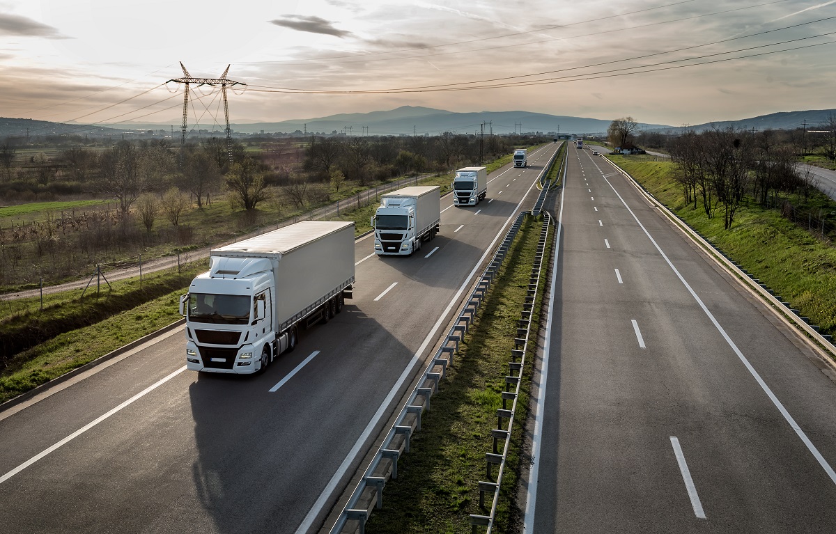 Aerial view of a wide highway with multiple white trucks traveling in the same direction. Lush fields and trees border the road, with mountains visible in the background under a cloudy sky. The scene conveys a sense of transport and movement.