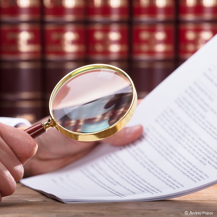A person holds a magnifying glass over a printed document, closely examining the text. Books with red covers form the background, suggesting a study or library setting. The image conveys a focus on detailed inspection and analysis of written material.