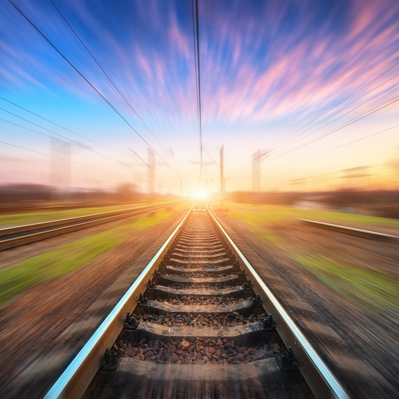 A view of a railway track stretching towards a sunset, with blurred colors creating a sense of motion. The rails are lined with gravel, and electrical poles are visible in the background, emphasizing the journey ahead in a tranquil yet dynamic atmosphere.