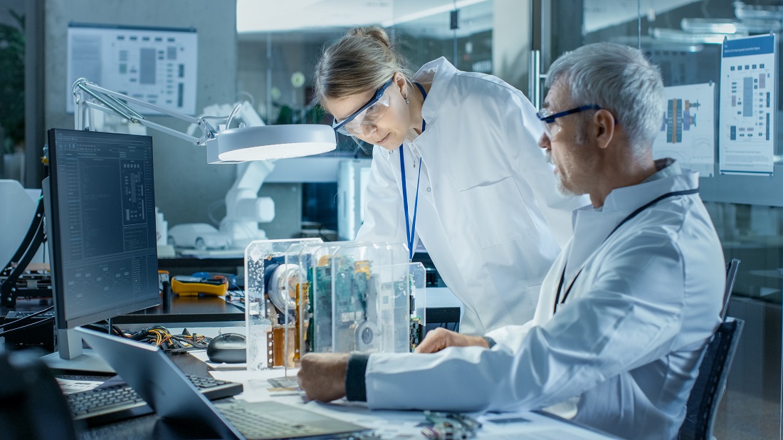 Zwei Laboranten in weißen Laborkitteln arbeiten an einem technischen Gerät. Eine Frau schaut konzentriert auf das Innere einer transparenten Box, während ein Mann ihr dabei assistiert. Beide sind von Computern und technischen Geräten umgeben.